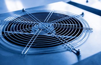 Close-up of a metal ventilation fan with a protective grille, mounted on a flat surface. The image has a blue tint, creating a cool and industrial appearance.