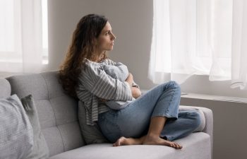 Woman sitting cross-legged on a sofa, looking out the window, holding a blanket.