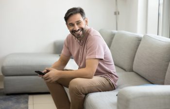 Middle-aged man sitting on couch with mobile phone, casually dressed resting on sofa alone at home, looking away with relaxed and cheerful expression, received good news, engaged in enjoyable chat