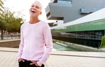Man in a pink sweater standing outside a modern building, smiling with hands in pockets.